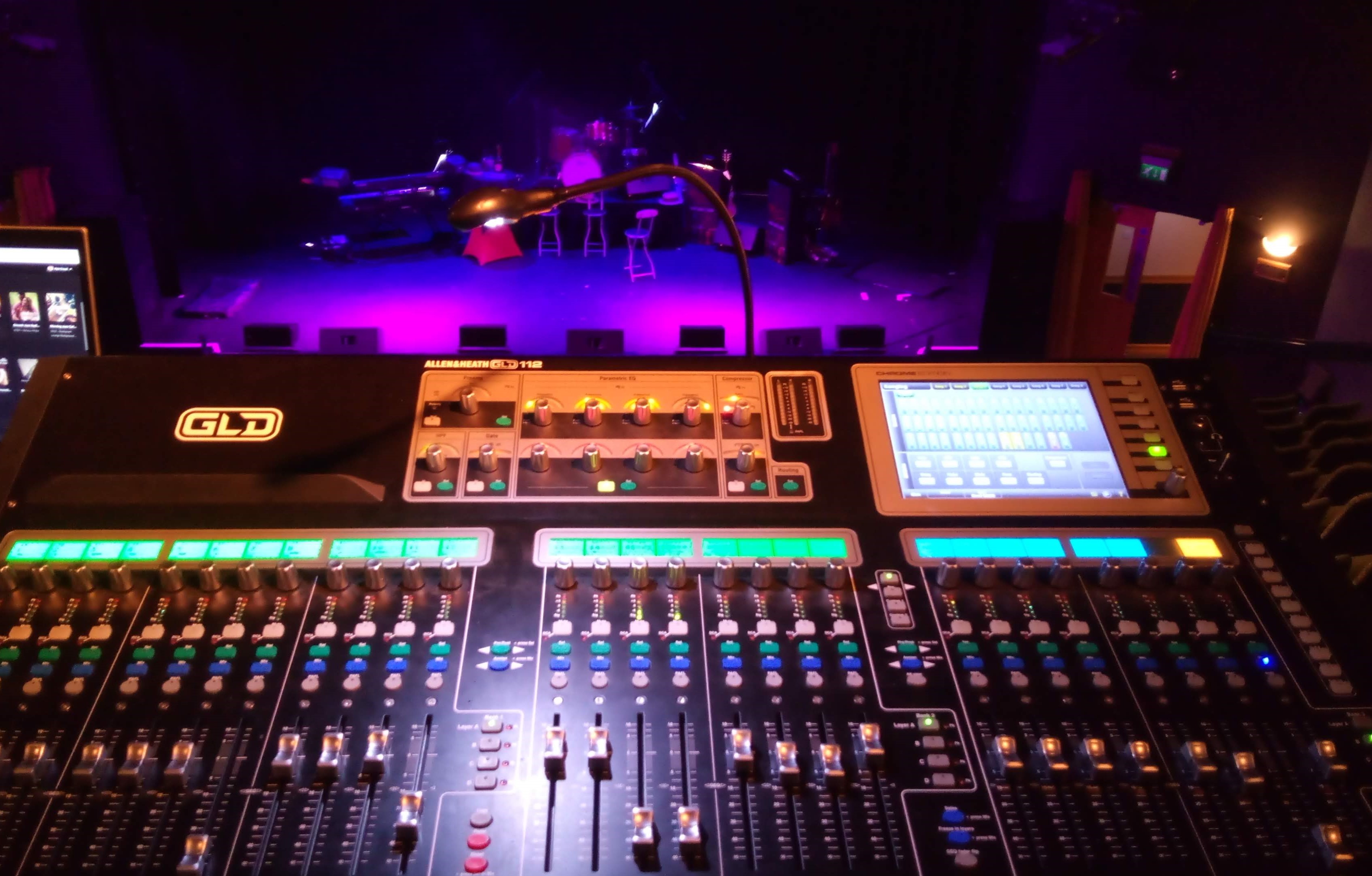 In the foreground, a large sound mixing desk with lots of sliders, buttons, knobs and screens.  In the background, a dimly lit stage: several bar stools and a large drum kit are just visible.