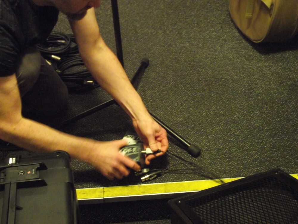 A man kneels betweem a microphone stand and a monitor speaker as he connects one plug of a multicore cable into a DI box.
