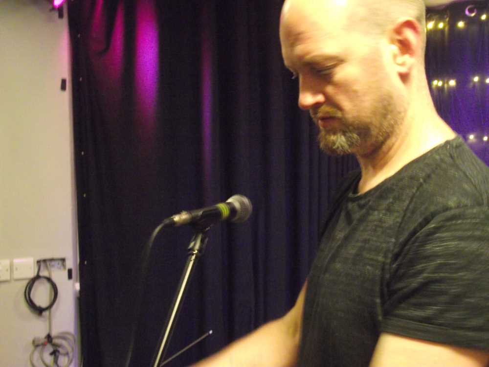 A man stands looking left, frowing in concentration at something we cannot see. In the background, a microhone sits on a stand, purple light shines on a black curtain, and a star cloth is just visible over the man's shoulder.