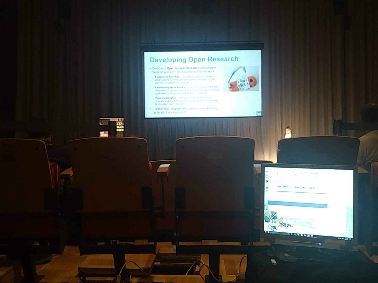 In the foreground, a computer monitor sits just in front of a row of chair backs. Over the chairs,  we can just see a figure at a lectern giving a talk, with their slides projected onto a large screen behind them. The slide title is clearly legible: 'Developing Open Research'