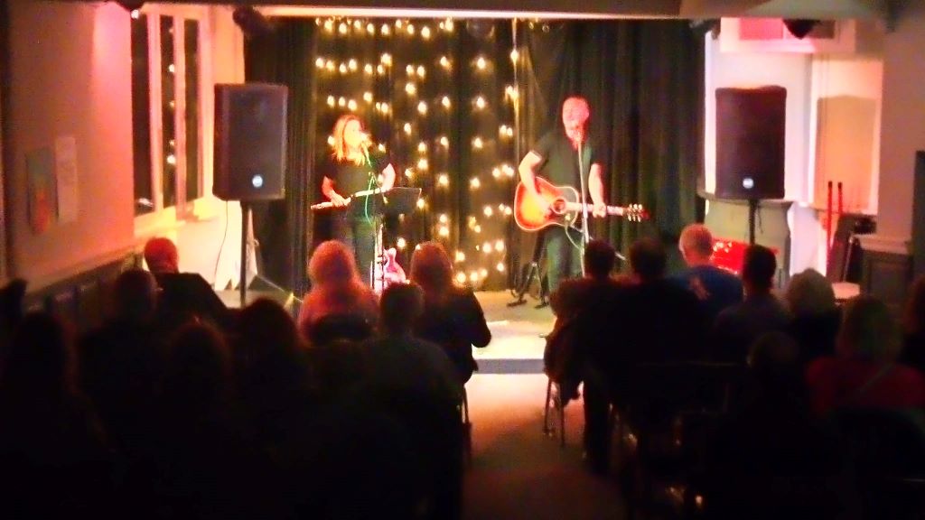In a blurry photograph, we look over the heads of a seated audience. Two people stand on stage in front of a star cloth;  the figure on the right is holding a guitar. They are flanked by large PA speakers.
